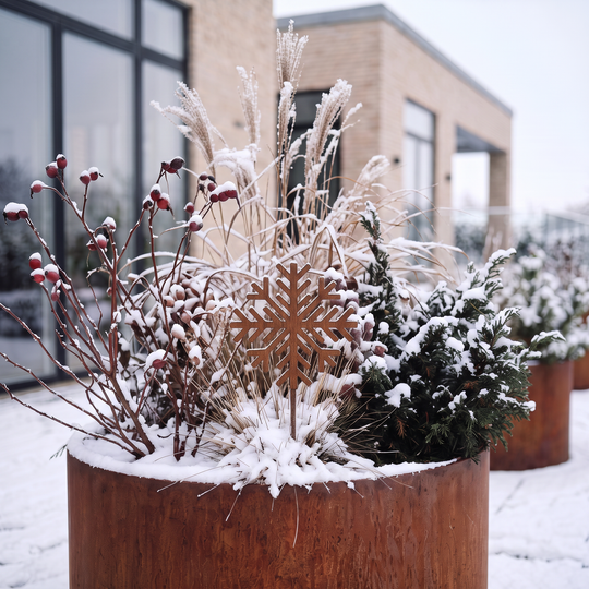 Gartenstecker in Form einer Schneeflocke aus Cortenstahl in einem runden, verschneiten Pflanzkübel mit Gräsern und Nadelgehölz.