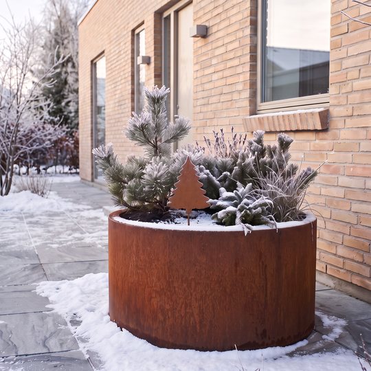 Gartenstecker in Tannenbaumform aus Cortenstahl in einem runden Pflanzkübel mit frostbedeckten Nadelgehölzen vor einer Backsteinfassade.