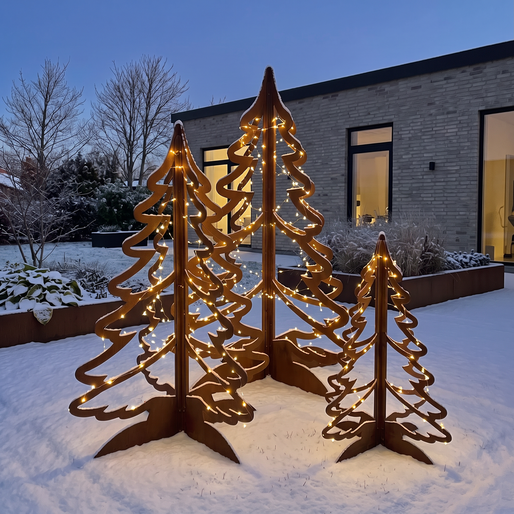 Drei beleuchtete Weihnachtsbäume aus Cortenstahl im verschneiten Garten in der Abenddämmerung vor einem modernen Haus.